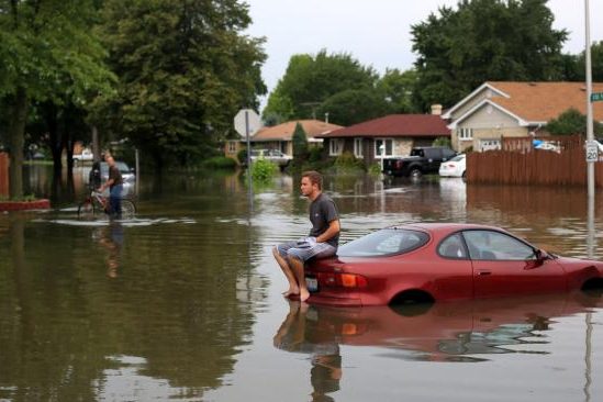 Een rode auto staat half onder water, met mensen die door een overstroming lopen.